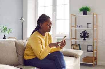 Young african american girl using mobile phone while sitting on sofa at home. Woman resting on couch with cellphone and looking at screen with worried face expression, chatting or typing sms message. - obrazy, fototapety, plakaty