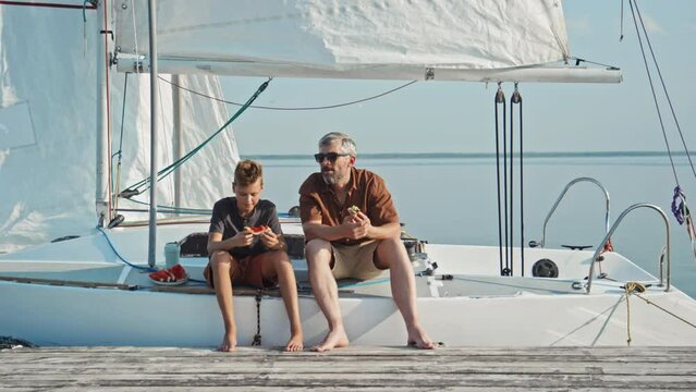 Long shot of teen boy and his dad sitting on yacht with their feet on wooden pier chatting about something while having lunch
