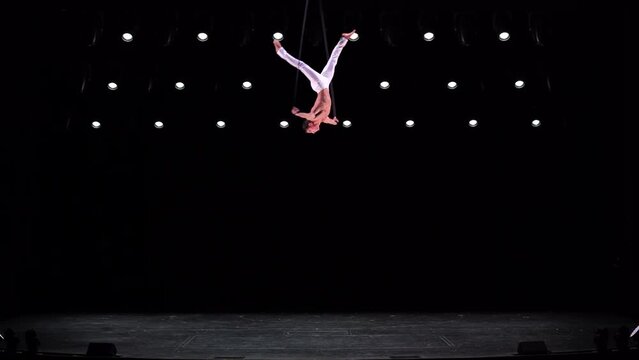 Male, muscular circus artist on Aerial straps on black stage background performing gymnastics trick