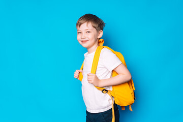Schoolboy with yellow backpack stands sideways against blue background.