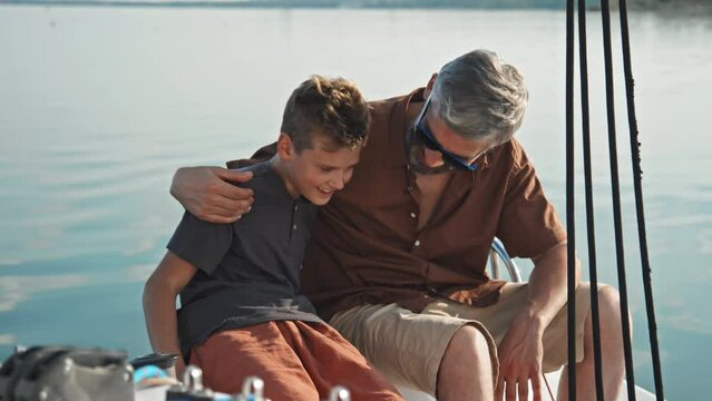 Happy mature Caucasian father sitting on board the sailing yacht enjoying spending summer day with his teen son