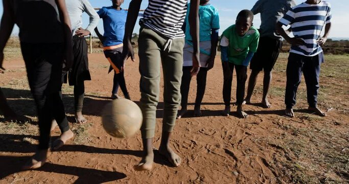 Poverty. Poor Black African child bouncing soccer ball on foot while team looks on in a township slum