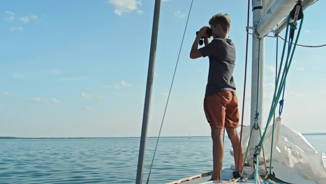 Long shot of adventurous teen boy spending time on sailboat looking into distance using binoculars