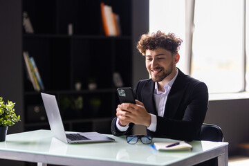 Smiling businessman chatting on phone in office. He is sitting at desktop with laptop