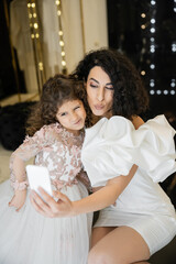 funny middle eastern bride with brunette hair in white wedding dress with puff sleeves and ruffles taking selfie on smartphone with daughter while pouting lips in bridal store