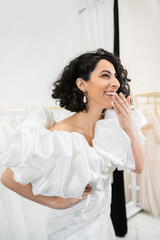radiant middle eastern bride with brunette wavy hair laughing, covering mouth and posing in trendy...