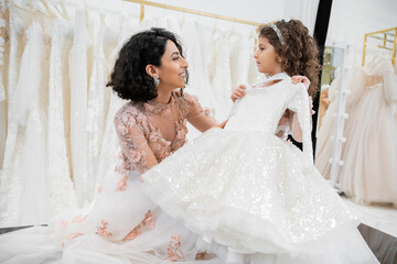 Naklejka premium happy and brunette middle eastern woman in floral wedding gown helping to choose white dress for her little daughter in bridal salon around tulle fabrics, process of preparation
