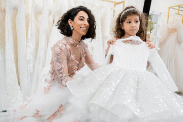 Naklejka premium cheerful and brunette middle eastern bride in floral wedding gown looking away near her little daughter with cute dress in bridal salon around white tulle fabrics, process of preparation