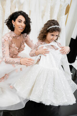 happy and brunette middle eastern bride in floral wedding gown helping to choose dress for her little daughter in bridal salon around white tulle fabrics, process of preparation