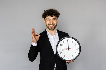 Laughing young bearded business man in classic black suit shirt tie posing isolated on grey background holding clock
