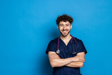 Happy smiling doctor or male nurse in blue uniform with crossed arms over blue background - obrazy, fototapety, plakaty