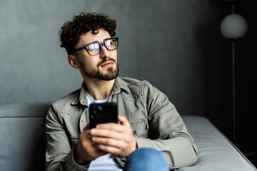 Thoughtful serious man looking at smartphone screen, sitting on couch at home. Young businessman or student reading bad news in message, unexpected debt, solving problem