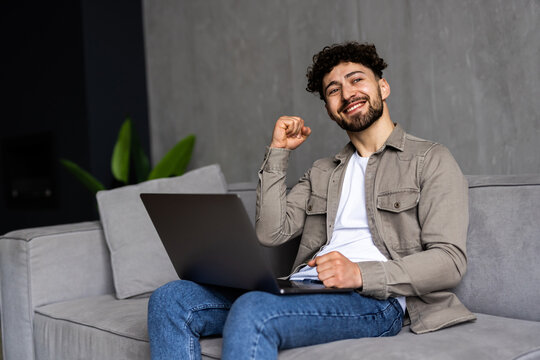 Smiling Young Man Holding Mobile Phone While Sitting On A Sofa At Home With Laptop And Celebrating