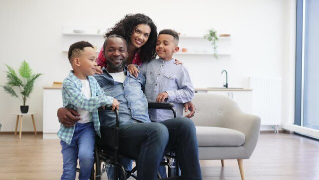 Full length portrait of happy multicultural family of four hugging each other while posing in kitchen of modern flat. Smiling mother cuddling two cheery kids and relaxed husband in wheelchair.