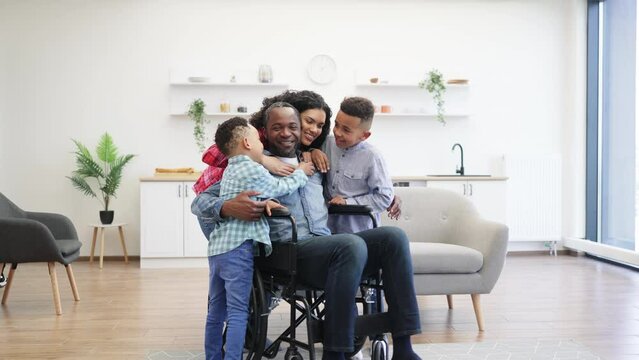 Full length view of african american parents showing their care while embracing smiling kids in studio apartment. Handsome father with disability telling jokes and making little boys laugh at noon.