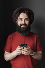 Portrait of handsome young man using his phone while standing against black wall. He is wearing an red t-shirt a black hat