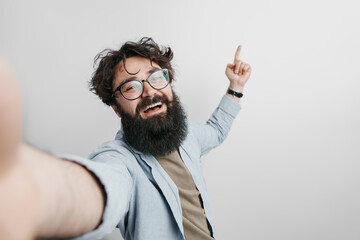 Joyful bearded man wearing eyeglasses and denim shirt taking selfie against white background