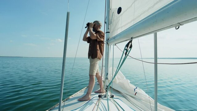 Long shot of mature Caucasian man standing barefoot on board of yacht looking into distance through binoculars