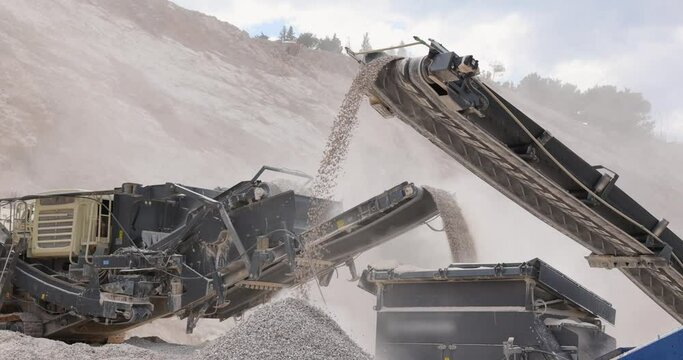 Stone crushing machine at open pit mining and processing plant for crushed stone. Piles of gravel stone and conveyor belts on the side of the road, slow motion