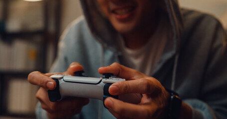 Close-up Happy young Asian man wear gray hoodie hold joystick controller excitement and shouting while having fun winning playing video game sit on sofa in the living room home at night. © tirachard