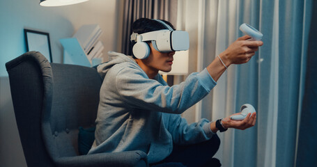 Young Asian man gamer wear gray hoodie with VR controllers headset hold joystick controller creating 3D art sit on sofa living room home at night. Futuristic artwork virtual reality concept. © tirachard