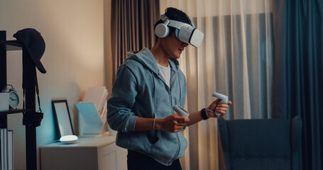 Young Asian man gamer wear gray hoodie with VR oculus hold VR joystick controllers feeling exciting with new experience of game stand front sofa living room home at night. Virtual reality concept. © tirachard