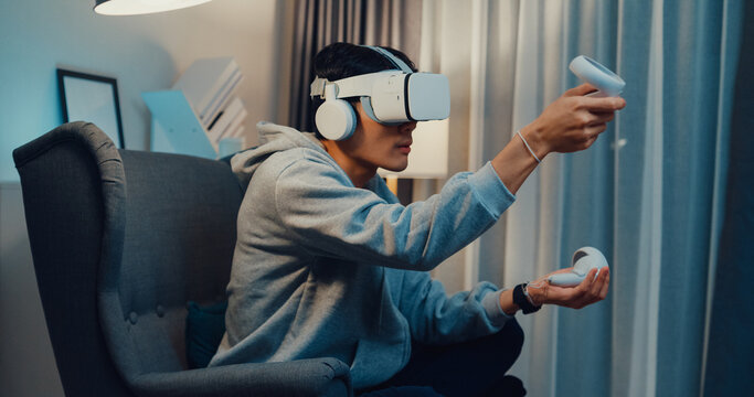 Young Asian man gamer wear gray hoodie with VR controllers headset hold joystick controller creating 3D art sit on sofa living room home at night. Futuristic artwork virtual reality concept. - Powered by Adobe