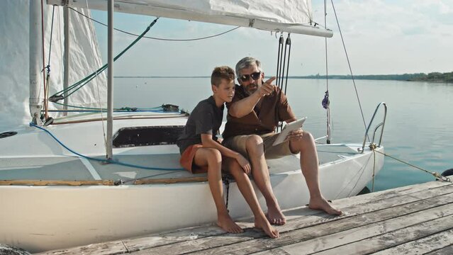 Mature Caucasian tourist and his teen son sitting on yacht with feet on pier reading map on digital tablet