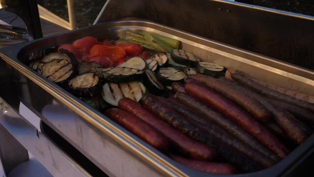 Female hand opens the lid of a pan in which vegetables are grilled sausages.