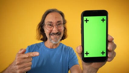 Excited mature elderly man standing and pointing Big smartphone with blank green screen with tracking points, demonstrating copy space for app or website design, mockup image on yellow background