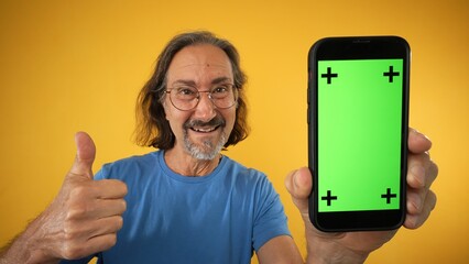 Excited mature elderly man standing and pointing Big smartphone with blank green screen with tracking points, demonstrating copy space for app or website design, mockup image on yellow background