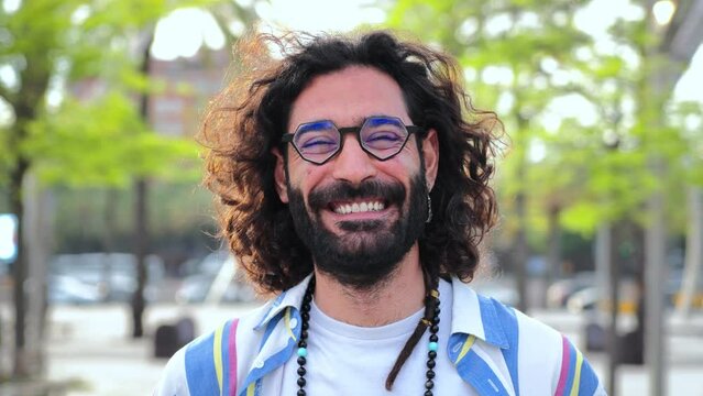 Happy young hipster man smiling and looking at camera. Close up individual portrait of hippie guy laughing. Front view of one bearded bohemian male person standing outdoors with cool expression. High