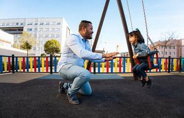 A Dominican father plays Happily with her daughter at an outdoor playground. The black man pushes his daughter from the front who is sitting on a swing. Concept of fun in playgrounds.