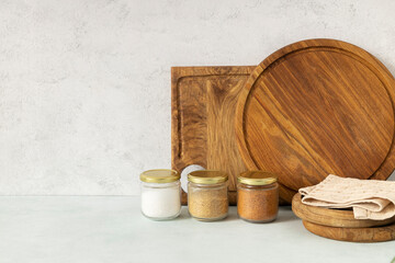 Fototapeta premium Kitchen table and wooden cutting boards, trays and glass jars for spices against white wall