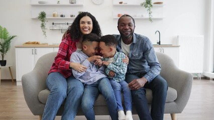 Full length view of african american couple with two boys posing on soft couch in living room of modern apartment. Loving mom and dad embracing little sons while spending quality time together.