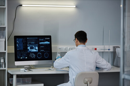 Back view at male scientist at desk in laboratory with data on computer screen, copy space