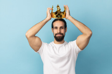 Portrait of serious independent man with beard in white T-shirt wearing golden crown, looking with...