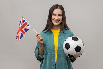 Happy positive woman holding in hands flag of United Kingdom and football ball, supporting favourite team on championship, wearing casual style jacket. Indoor studio shot isolated on gray background.