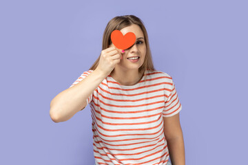 Portrait of positive funny young adult blond woman wearing striped T-shirt holding little red heart and covering her eye behind love symbol. Indoor studio shot isolated on purple background.