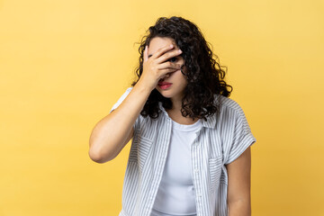 Portrait of curious nosy woman with dark wavy hair spying through hole in fingers closing eyes with arm, peeking, sees something interesting. Indoor studio shot isolated on yellow background.