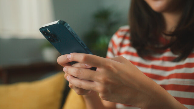 Close up of beautiful Asian woman sit on sofa hand holding mobile phone chatting with friends and playing social media. Young female looking at smartphone cellphone browsing internet at living room