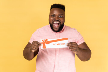 Portrait of excited amazed happy man wearing pink shirt holding gift voucher, looking at camera...