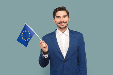 Portrait of smiling happy handsome man with mustache standing waving europe union flag, expressing happiness, wearing white shirt and jacket. Indoor studio shot isolated on light blue background.