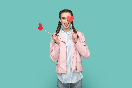 Portrait of romantic positive teenager girl with braids wearing pink jacket standing covering eye with red heart on stick, showing tongue out. Indoor studio shot isolated on green background.