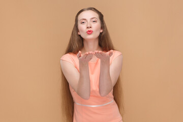 Portrait of romantic flirting pleased attractive woman with long hair sending air kissing, expressing her devotion, wearing elegant dress. Indoor studio shot isolated on brown background.