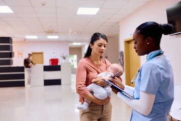 Caring mother with baby talks to pediatric nurse in waiting room at clinic.