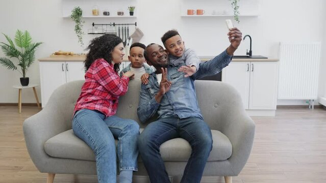 Positive multicultural family of four using modern smartphone while resting in living room. Adorable sons embracing parents while dad taking selfie via mobile webcam in room interior.