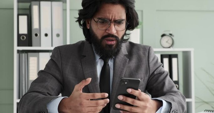 An Indian businessman in glasses and a suit with a tie is sitting at his desk, texting on his phone. Suddenly, he sees something on the screen that upsets him and he loses his temper.