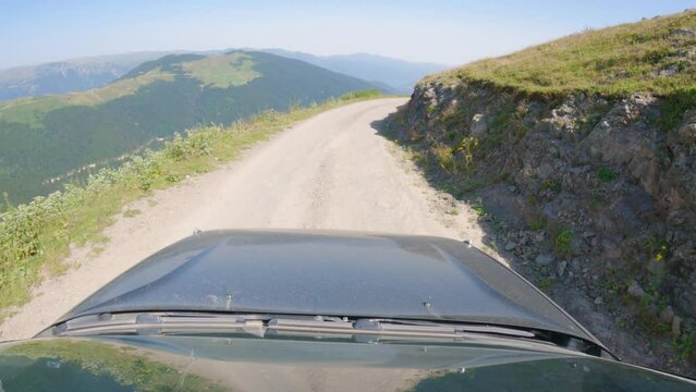 The car is driving on a narrow dirt road. A dangerous road in mountains. View from roof of car, with bonnet in frame. Car driving on a mountain road. Beautiful mountain scenery in background