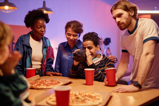 Portrait of ethnic young man with retro video camera filming friends eating pizza at house party